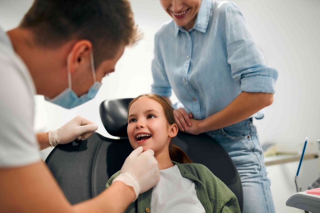 Dentist examining a smiling child’s teeth while a parent looks on.