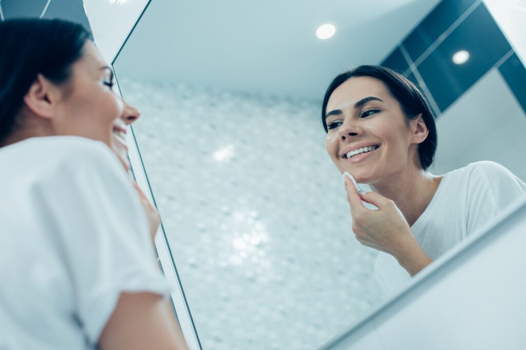 contented-young-lady-smiling-looking-mirror-cleaning-her-face-with-cotton-pad